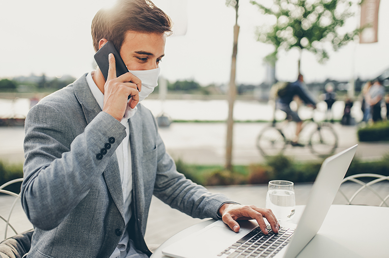 A man works outdoors on the phone and a laptop wearing a surgical mask